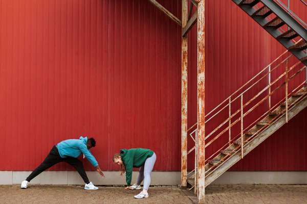 Two people stretching beside red metal building and stairs.