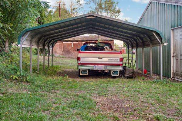 a metal carport sheltering a pickup truck, with grass and trees around the area